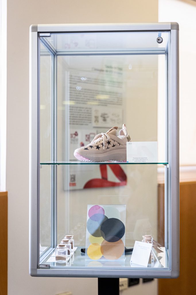 Teca in vetro contenente una scarpa sportiva in pelle di kiwi e sul ripiano inferiore dei campioni di pellami realizzati con la buccia della frutta