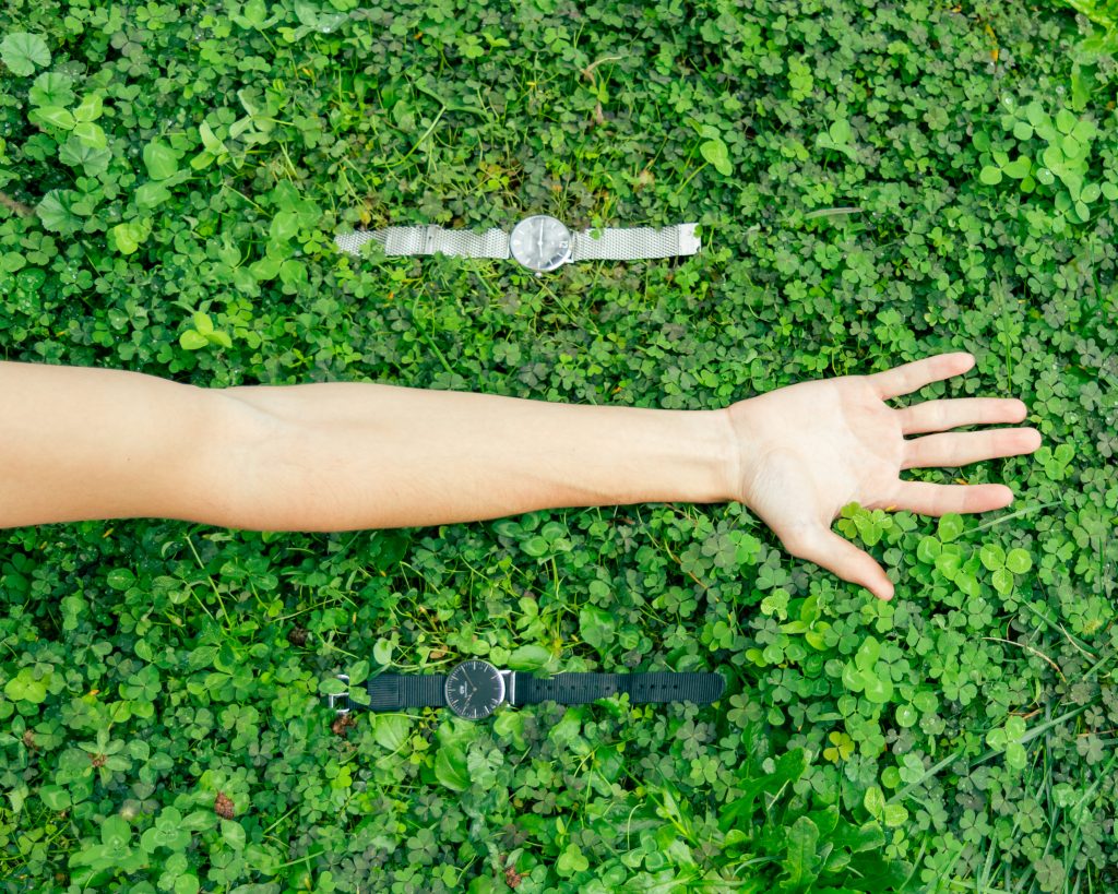 Fotografia che mostra il braccio di un rgazzo con la mano rivolta verso l'alto, appoggiato su di un prato. Accanto al braccio ci sono un orologio grigio in alto e uno nero in basso