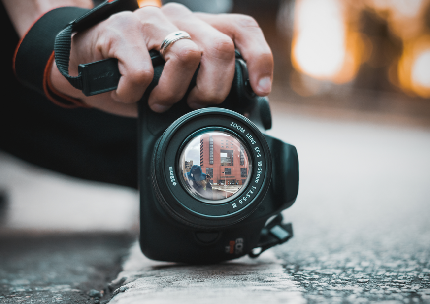 Immagine raffigurante una mano che sostiene una macchina fotografica. Nell'obiettivo si vede una persona che scatta una foto all'edificio U12